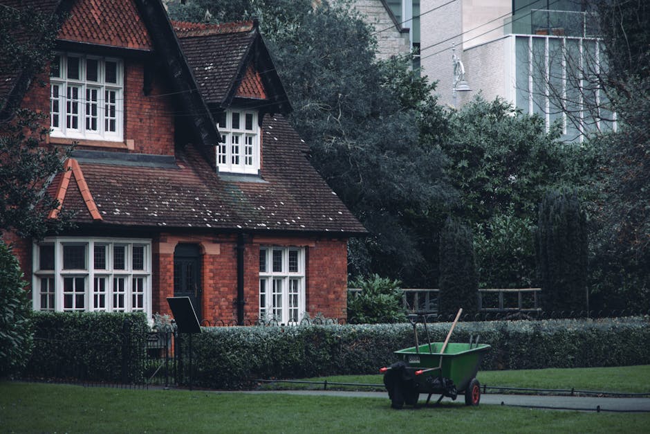 A residential garden scene featuring a well-maintained lawn with lush green grass and neatly trimmed hedges along the edges. The garden includes a low brick wall and a black wrought-iron fence surroun