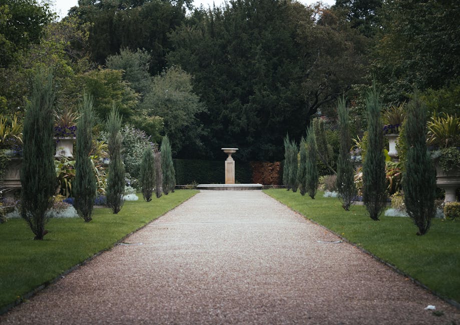 A gravel pathway in a well-maintained garden leads straight ahead towards a classical pedestal fountain in the distance, framed by evenly spaced, tall, narrow cypress trees on both sides. The pathway 
